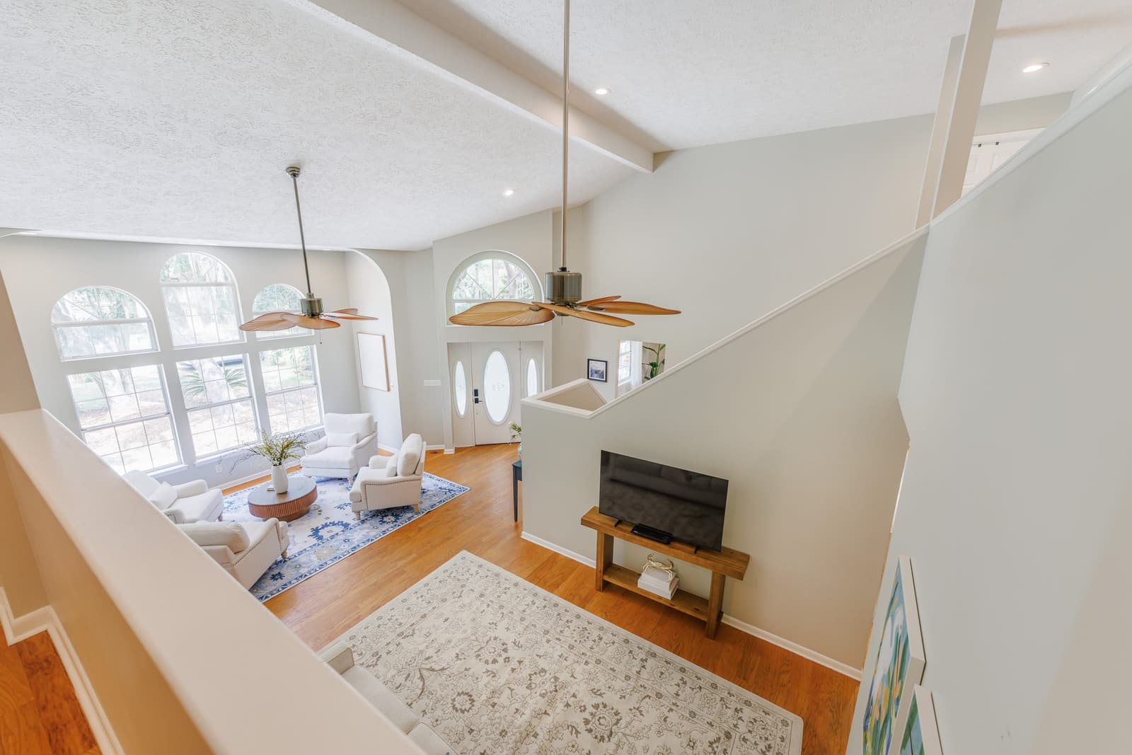 View across the living room from the stairs, with ceiling fans, arched windows, and the TV area